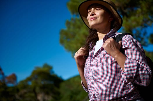 A confident woman enjoys hiking in a sunny outdoor landscape. She wears a hat and backpack, looking content and adventurous. The blue sky adds to the tranquil setting.