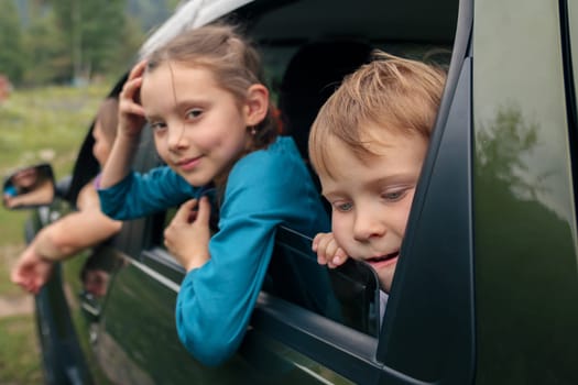 Two cheerful children happily peek out from the car window, thoroughly enjoying a vibrant outdoor adventure