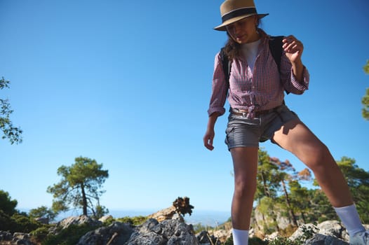 A young woman wearing a hat and carrying a backpack hikes through a rocky landscape. She is joyfully exploring a mountainous area on a sunny day, capturing the essence of adventure.