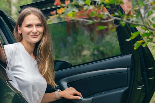 A young woman stands by her car, smiling broadly, amidst vibrant greenery and enjoying a beautiful moment outdoors