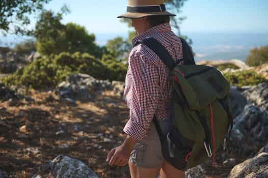 A woman hikes through a rocky trail surrounded by natural scenery, wearing a backpack and sunhat. It's a bright, sunny day ideal for outdoor exploration and adventure in nature.