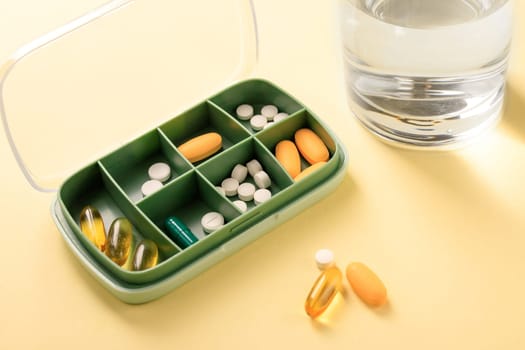 Green pillbox filled with a few types of pills and capsules on yellow background and a glass of water standing near.