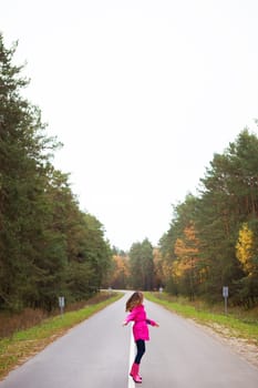 a girl in a pink raincoat and rubber boots turns on the road in the middle of the forest.