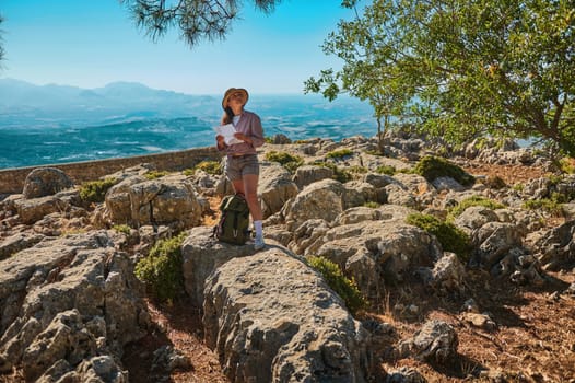 A young woman hiker stands amidst rocky terrain, holding a map and wearing a backpack, under clear blue skies. She appears to be exploring or navigating the natural landscape.