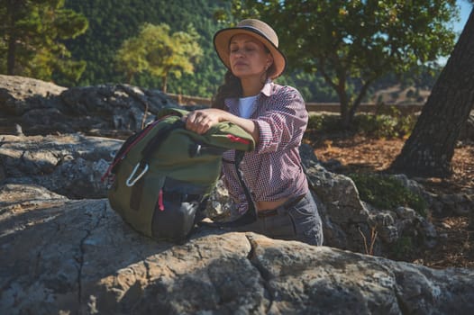 Woman enjoying a peaceful hiking break in lush nature, packing her backpack on rocky terrain.