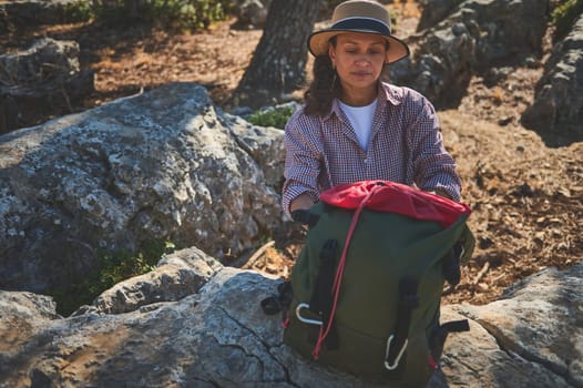 A woman in a hat and checked shirt prepares her green backpack on rocky terrain. Surrounded by nature, she embodies the spirit of adventure and exploration, enjoying a peaceful hiking experience.