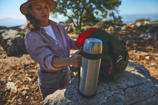 A woman takes a break during a scenic mountain hike, holding a thermos beside her backpack. The peaceful outdoor setting highlights a love for nature and adventure.
