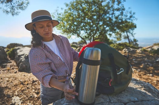 A woman dressed in a hat and checkered shirt prepares for a hike, standing beside a backpack with a thermos. The scenic mountain view and sunny weather enhance the outdoor adventure.