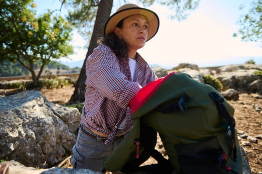 A woman in a hat and checkered shirt explores the outdoors with her backpack, surrounded by rocks and trees. The image captures the essence of adventure and connection with nature.