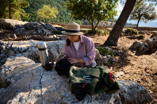 A woman is sitting on a rock, preparing a meal outdoors in a scenic park with lush greenery. She is dressed casually, surrounded by nature, embracing a moment of relaxation and leisure.
