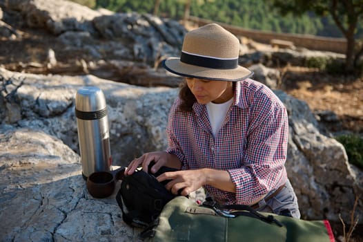 A woman spends time outdoors, sitting on rocky terrain with a backpack and thermos beside her. She is dressed casually in a hat and plaid shirt, preparing for a hike or adventure.