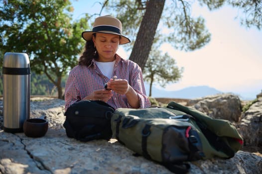 A woman sits on a rocky surface surrounded by nature, enjoying a serene outdoor break. She is checking her phone, with travel essentials and a thermos by her side in a peaceful setting.