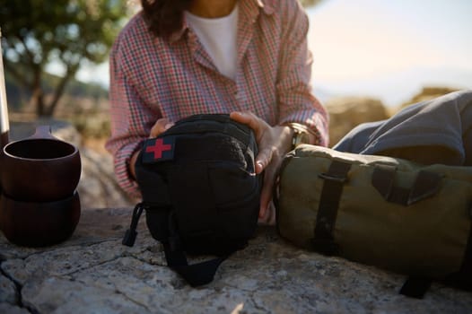 A woman sits on rocks organizing a first aid kit, surrounded by camping gear, highlighting outdoor preparedness. Sunlit and natural, it represents adventure and safety in nature.