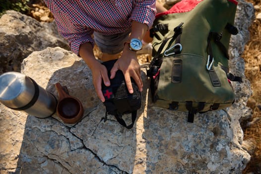 Top view of a hiker organizing a first aid kit on rocky terrain, with a backpack, thermos, and cup, highlighting outdoor adventure and readiness.