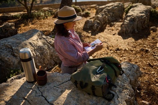 A woman wearing a hat reads a map while sitting on rocks in a sunny, natural setting. A backpack and thermos sit beside her, suggesting a day of adventure and exploration.