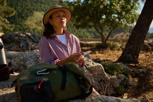 A woman in a hat enjoys a tranquil moment sitting on rocks surrounded by nature. She holds a paper, with a backpack and flask by her side, embodying relaxation and adventure.