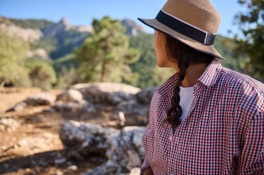 A woman wearing a straw hat and checkered shirt gazes at lush green mountains, capturing the essence of outdoor exploration and tranquility.