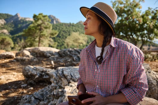 A woman in a straw hat enjoys a tranquil moment in nature, surrounded by mountains and greenery, evoking relaxation and exploration.
