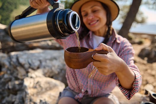 A cheerful woman pours a hot drink from a thermos into a wooden cup while enjoying the outdoors. The sunlight and natural setting suggest relaxation and exploration, embodying adventure.