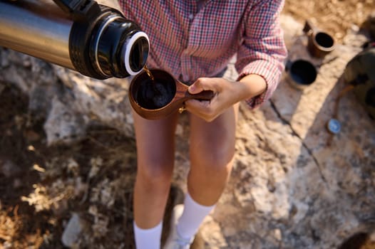 A person pours coffee from a thermos into a cup while sitting on rocky ground during a hiking trip. Casual clothing, nature, and relaxation are featured, capturing the essence of outdoor adventure.