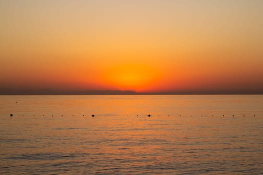 An orange sunrise lights up the sea, with the glowing sun rising over the horizon. The calm ocean reflects the vibrant colors, creating a serene and breathtaking view
