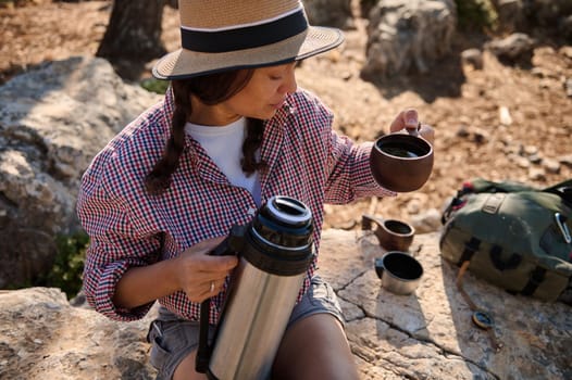 A woman wearing a hat and checkered shirt pours a hot drink from a thermos during a hiking trip. She sits on rocky terrain, exemplifying a peaceful outdoor adventure.