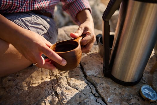A person's hands holding a wooden cup filled with a hot beverage, sitting beside a thermos on rocky terrain. The scene embodies tranquility, warmth, and the spirit of outdoor adventure.