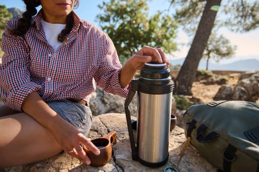 A woman enjoys a peaceful outdoor picnic on a sunny day, sitting on rocky terrain with a thermos and cup. Trees and mountains are visible in the background, creating a serene atmosphere.
