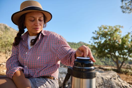 A woman sitting outdoors, wearing a hat and checkered shirt, enjoys a calm moment with a thermos. The natural setting, warm sunlight, and relaxed posture convey tranquility and leisure.