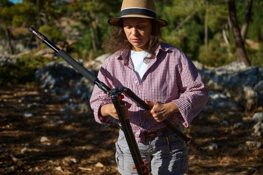 Woman in casual outdoor attire setting up a tripod against a natural park background during sunny day.