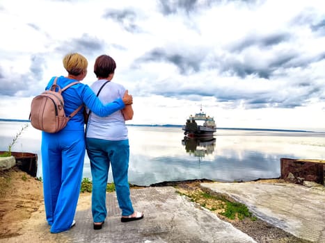 Two women stand together, watching a boat reflected on the water while enjoying a cloudy day.