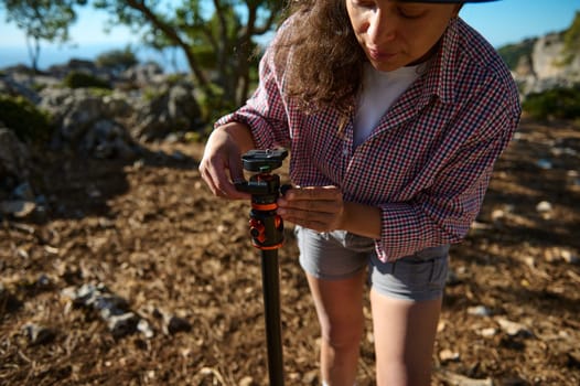 A woman is seen adjusting a camera tripod in a rustic outdoor environment. She is wearing casual attire, capturing the essence of natural photography. The scene is set in a sunlit, wooded area.