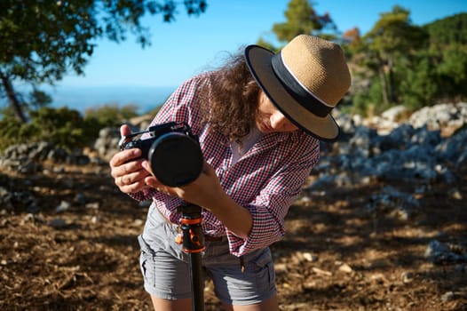 A woman adjusting her camera on a tripod while enjoying a photography session outdoors, surrounded by natural scenery and sunlight. She wears a straw hat and a checkered shirt.