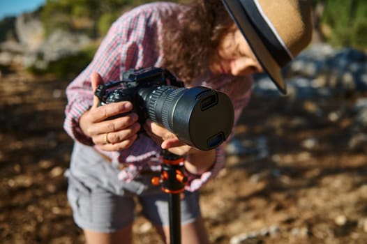 Close-up of a woman in a hat adjusting a camera on a tripod in a scenic outdoor environment, capturing the beauty of nature.