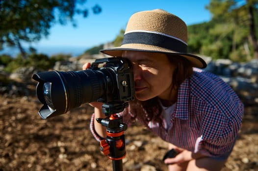 A woman wearing a straw hat and checkered shirt uses a camera mounted on a tripod to photograph nature in a sunny outdoor setting. The background is a scenic natural landscape.