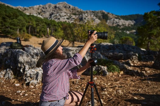 A woman in a hat and casual outfit prepares her camera on a tripod amidst a rugged mountain setting, capturing the essence of outdoor photography and exploration.