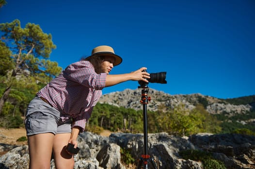A person sets up a camera on a tripod in a scenic outdoor location, surrounded by rocky landscapes and trees under a clear blue sky, capturing nature's beauty.