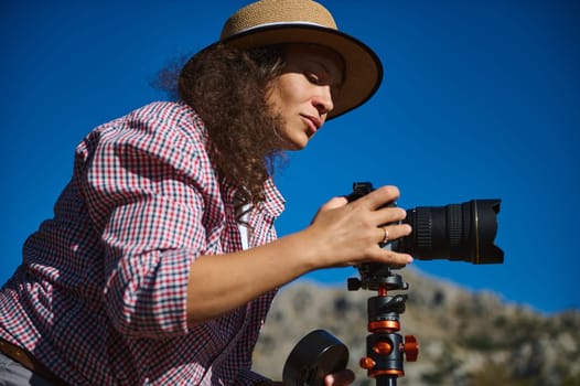 A dedicated photographer in a straw hat adjusts her camera settings, capturing the beauty of outdoor landscapes on a sunny day. Her concentration highlights her passion for photography.
