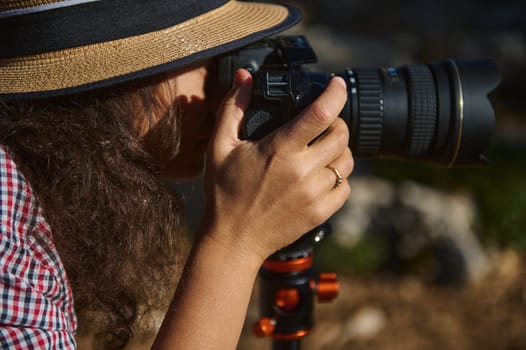 Close-up of a woman focusing through a DSLR camera lens, capturing outdoor scenes. Her hat and checked shirt add a casual, nature-loving vibe. Nature photography at its finest.