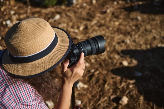 A photographer wearing a straw hat focuses on capturing outdoor scenery with a camera, creating an artistic composition in natural sunlight.