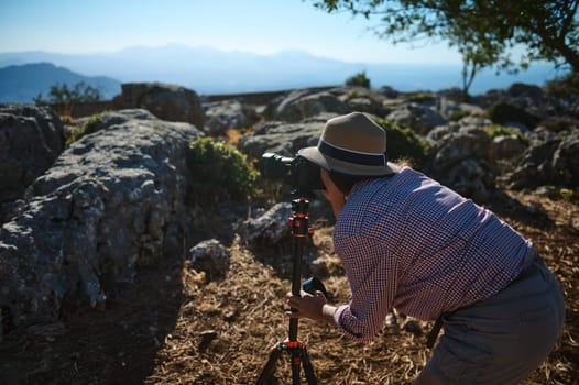 A man in casual attire uses a camera on a tripod to capture nature in a rocky terrain. The scene conveys exploration and a passion for photography amidst natural beauty.