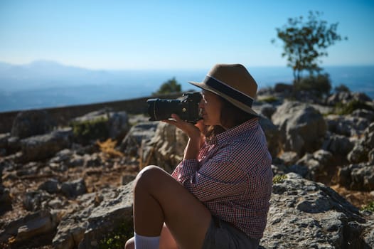 A woman captures the scenic view with her camera while sitting on rocky terrain under a clear sky. Her hat and relaxed posture reflect a peaceful and adventurous outdoor experience.