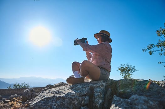 A woman sits on rocky terrain, using binoculars under a clear blue sky. The scene reflects adventure, exploration, and appreciation of nature's beauty.