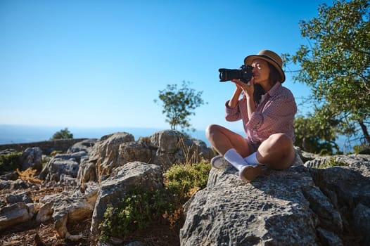 A woman sits on rocky terrain, capturing the scenic beauty through her camera, surrounded by nature. Her relaxed posture and focus embody the essence of adventure and exploration.