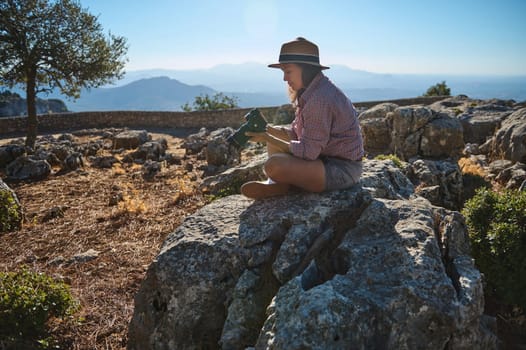 A woman in outdoor attire sits on a rock, exploring her surroundings with a camera. The landscape is rugged and scenic, providing a serene, natural atmosphere under clear blue skies.