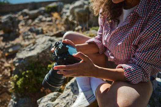 A woman in a plaid shirt sits on rocky terrain, adjusting her camera settings. The scene captures exploration, photography, and leisure in a natural environment.