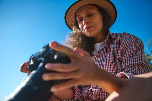 A woman wearing a straw hat and a plaid shirt examines her camera outdoors. The blue sky and natural light create a vibrant setting, symbolizing adventure and creativity.