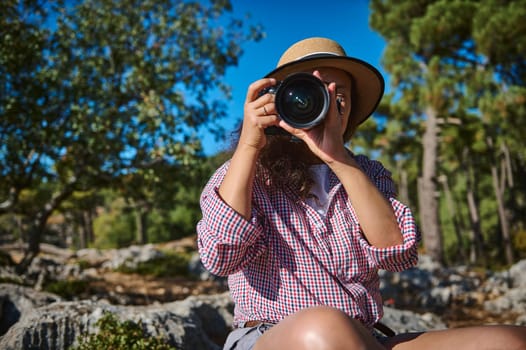 A woman wearing a hat holds a camera, capturing moments in a lush forest. The bright day highlights her passion for photography amidst nature. An image of exploration and creativity.