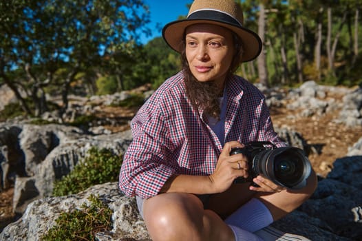 A relaxed woman with a camera sits amongst rocks and trees, enjoying the beauty of nature. Wearing a hat and casual clothing, she embodies the spirit of outdoor exploration and creativity.