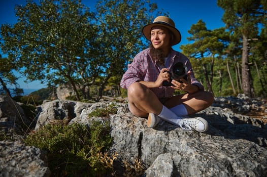 A woman sits on rocky terrain, surrounded by trees, holding a camera. She's wearing casual attire and a straw hat, enjoying the sunny day, capturing the essence of nature and photography.
